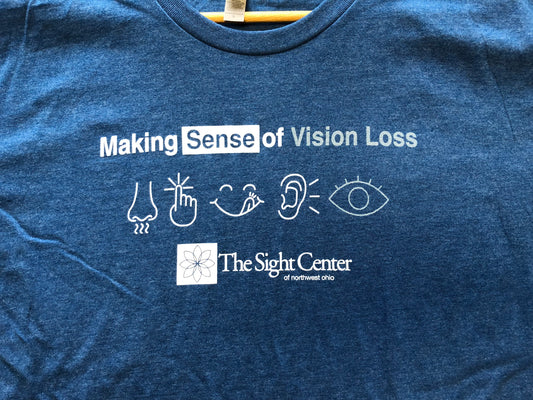 A blue t-shirt with white pictures and letters. It says Making Sense of Vision Loss. There are 5 pictures that represent the 5 senses. Smell, Touch, Taste, Hear and Sight. Below the pictures is the Sight Center of Northwest Ohio logo,