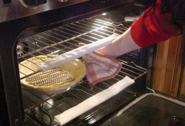 A hand with a pot holder reaches into an oven for a pie crust. Their aim is off and if weren't for the white oven rack bumpers they would have a nasty burn mark on their forearm.
