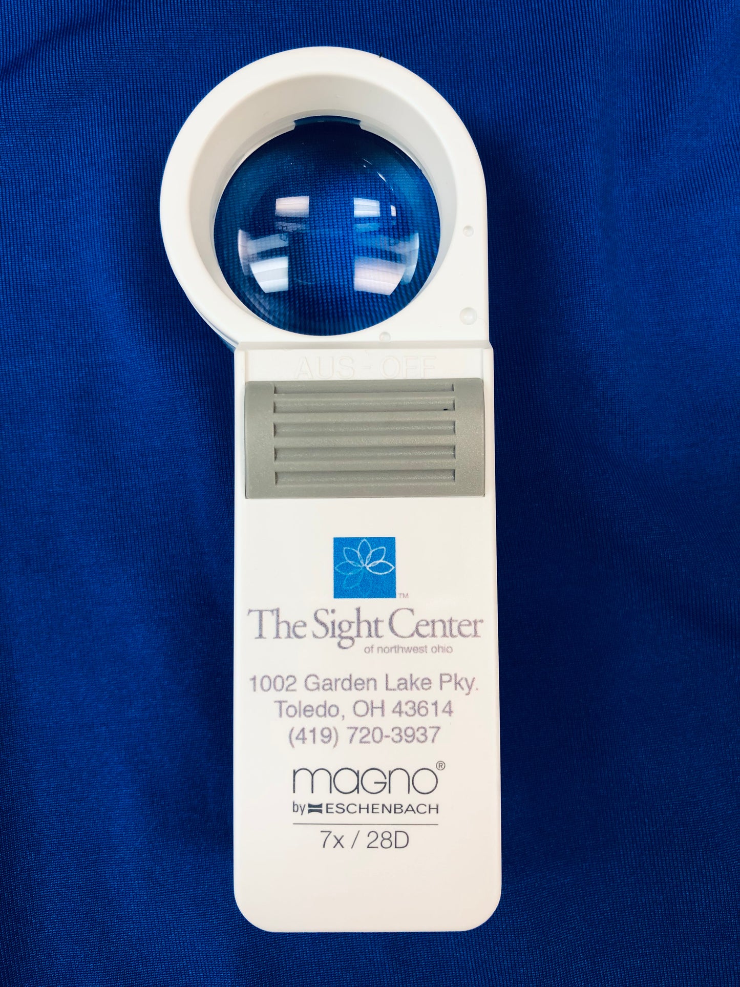 The 7x Magnifier has a circular viewing area. It features a grey on/off button. The Sight Center logo and contact information is printed on the handle along with the brand, Eschenbach, and the strength.