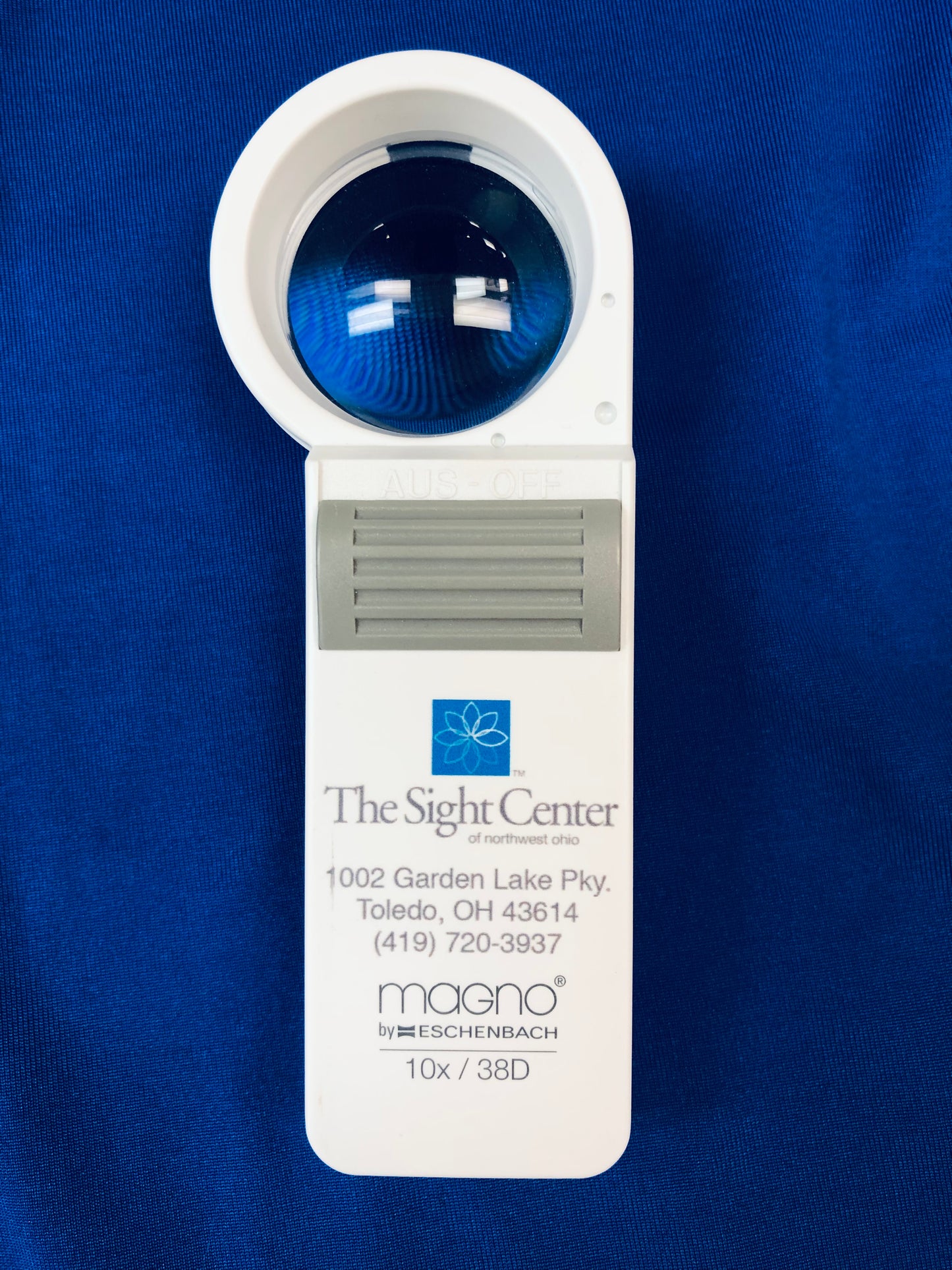 The 10x Magnifier has a circular viewing area. It features a grey on/off button. The Sight Center logo and contact information is printed on the handle along with the brand, Eschenbach, and the strength.