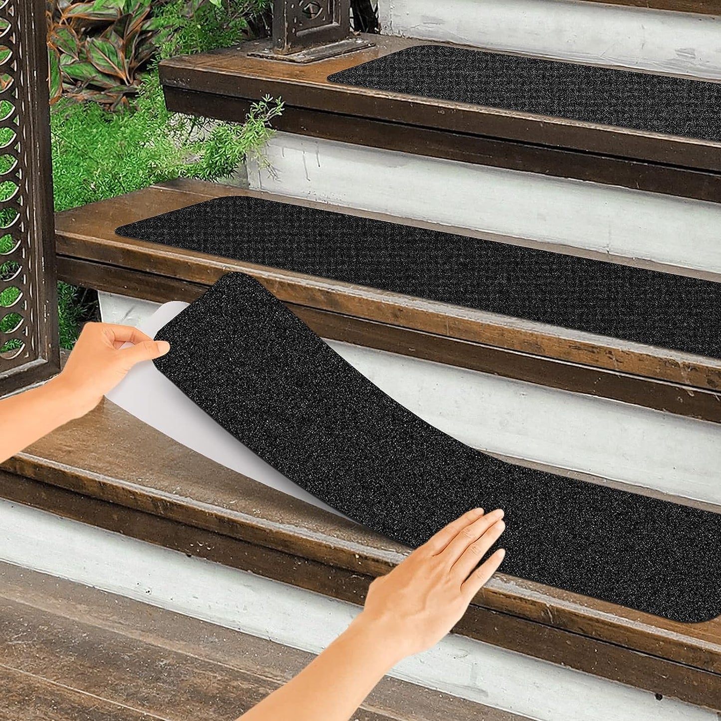 A pair of hands are placing black traction strips on a set of wooden stairs outside.